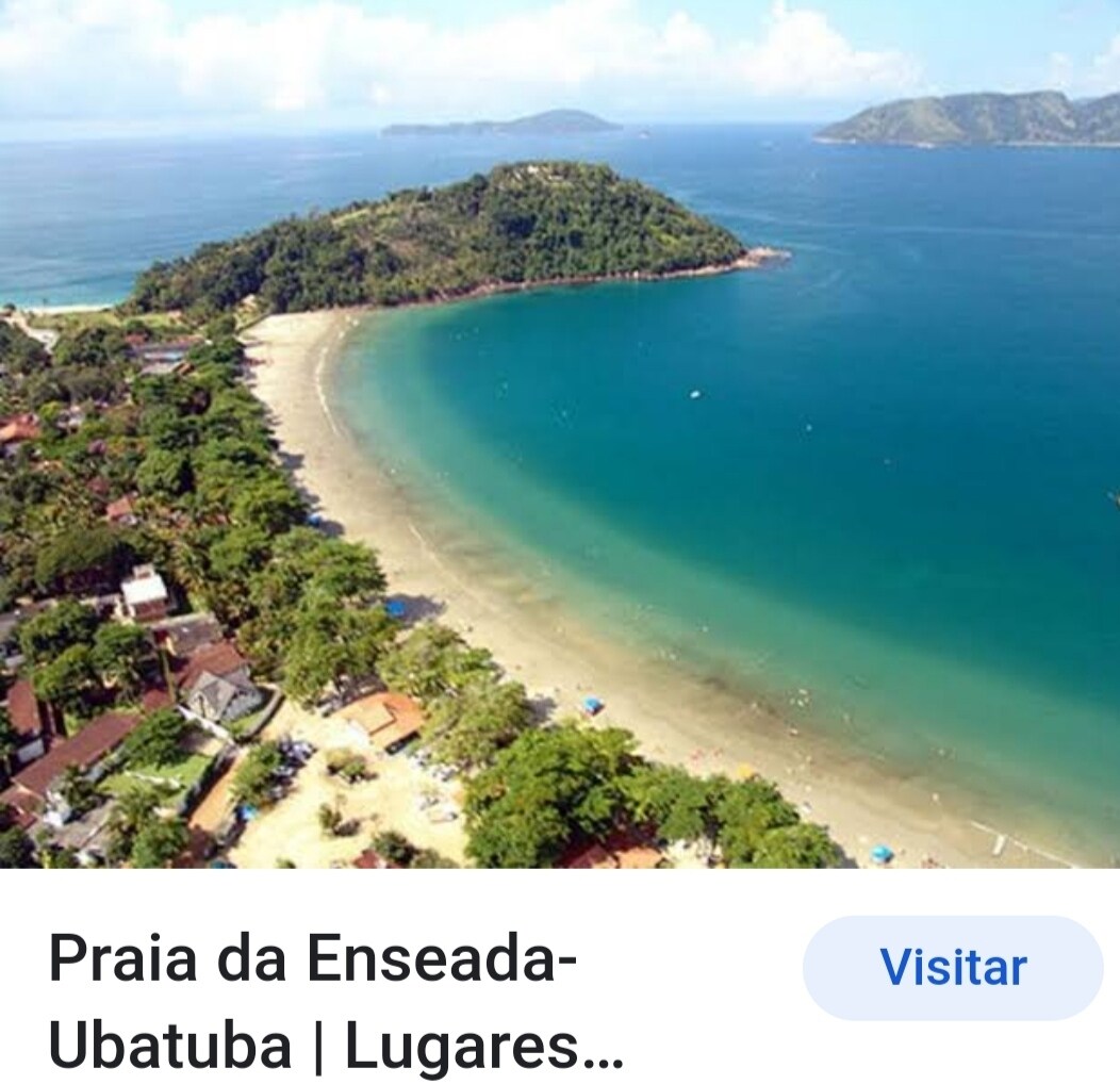Aerial view of a serene beach with soft, golden sand and gentle waves lapping at the shore. Lush greenery surrounds the shoreline, complemented by distant hills. The tranquil blue water is dotted with small boats, offering a glimpse of the vibrant coastal landscape.