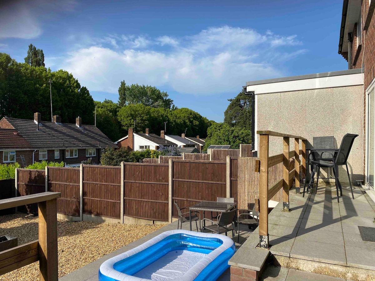 The image features a balcony view overlooking a garden area with a small inflatable pool. A wooden railing is visible, alongside garden furniture including a table and chairs. Behind the property, trees and neighboring houses are visible under a clear blue sky.