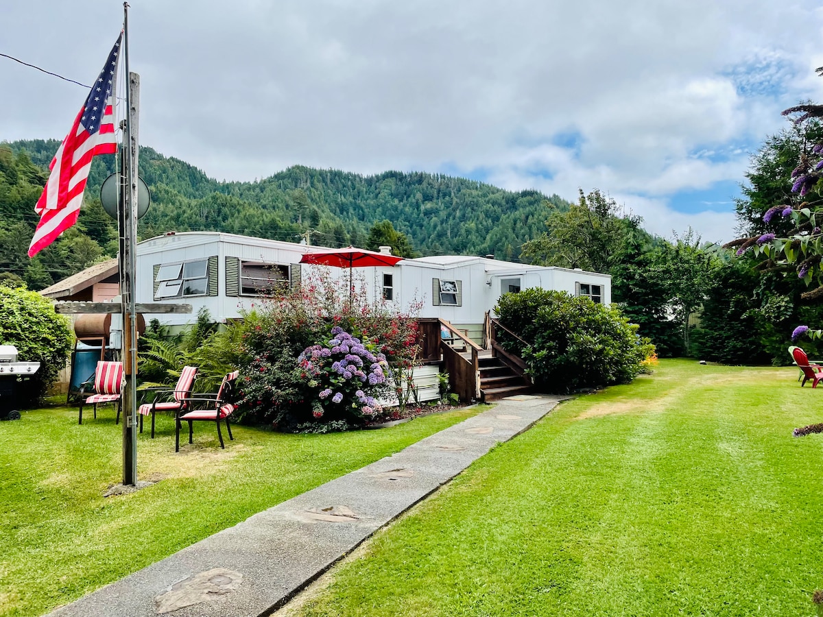 A well-maintained mobile home is visible, accompanied by vibrant purple flowers and neatly trimmed grass. A stone pathway leads to the entrance, with red and white striped chairs arranged on either side, and an American flag gently waving in the breeze.