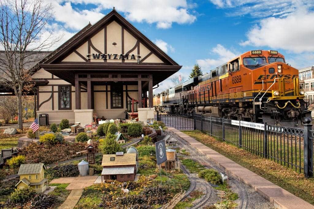 A charming train station is depicted, featuring traditional architecture with decorative elements. A train passes by, creating a sense of movement. Flower beds and low greenery surround the station, adding vibrant colors to the scene. A small model house is visible in the foreground.
