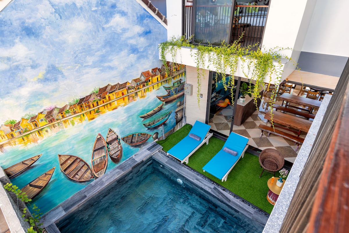 A swimming pool is complemented by a vibrant mural depicting traditional boats and houses by the water. Lounge chairs in a serene blue are positioned along the poolside, surrounded by greenery and a spacious patio area that invites relaxation.