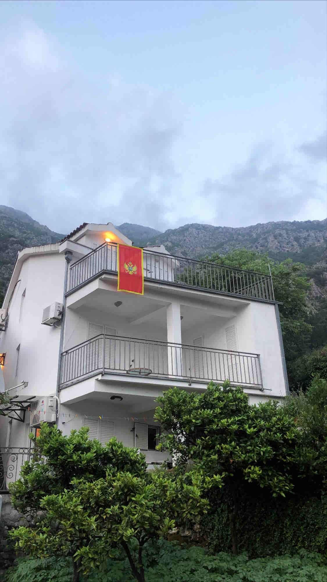 A modern three-story villa is visible, featuring multiple balconies with metal railings. The Montenegrin flag is displayed prominently on the upper balcony. Lush greenery surrounds the entrance, with mountainous terrain visible in the background under a cloudy sky.