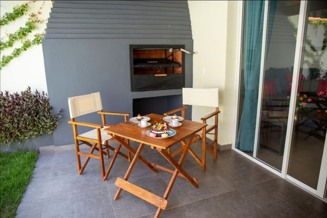 A cozy outdoor seating area features two folding chairs and a small wooden table set with dishes. A built-in barbecue grill is integrated into the wall, while lush greenery lines the space, creating a relaxing environment.