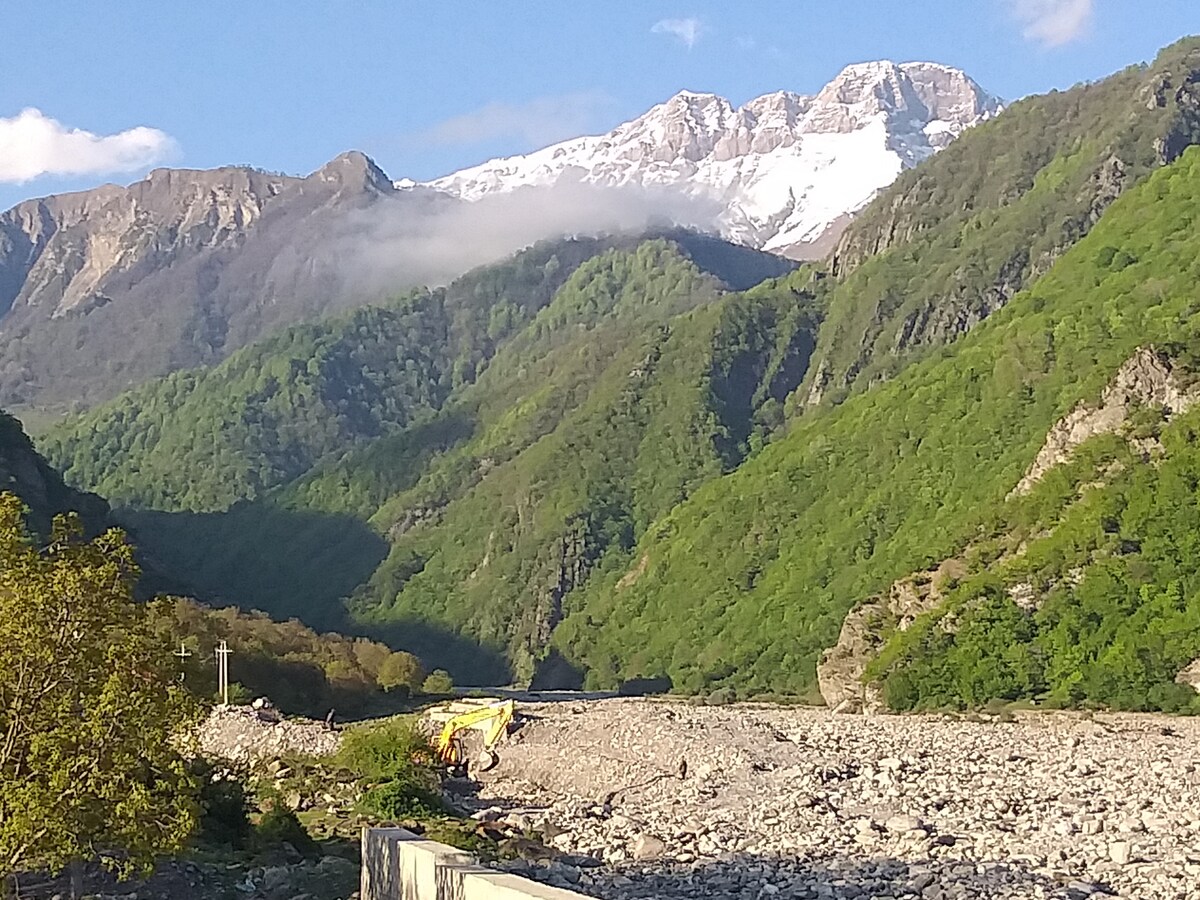 A scenic landscape is displayed, featuring lush green mountains and a snow-capped peak in the background. A riverbed with smooth stones runs through the foreground, leading towards the mountains. Gentle clouds hover near the summit, enhancing the natural beauty of the scene.