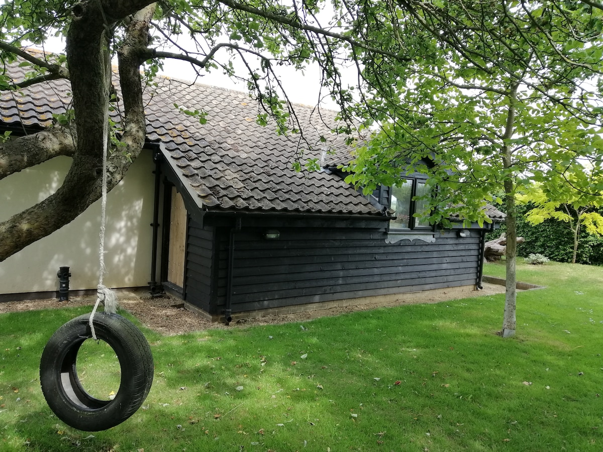 A charming black wooden exterior of The Garden Barn is complemented by a grassy area and trees. A swing made from a tire hangs from a tree branch, adding a playful element to the peaceful rural setting. Windows allow natural light to brighten the space.