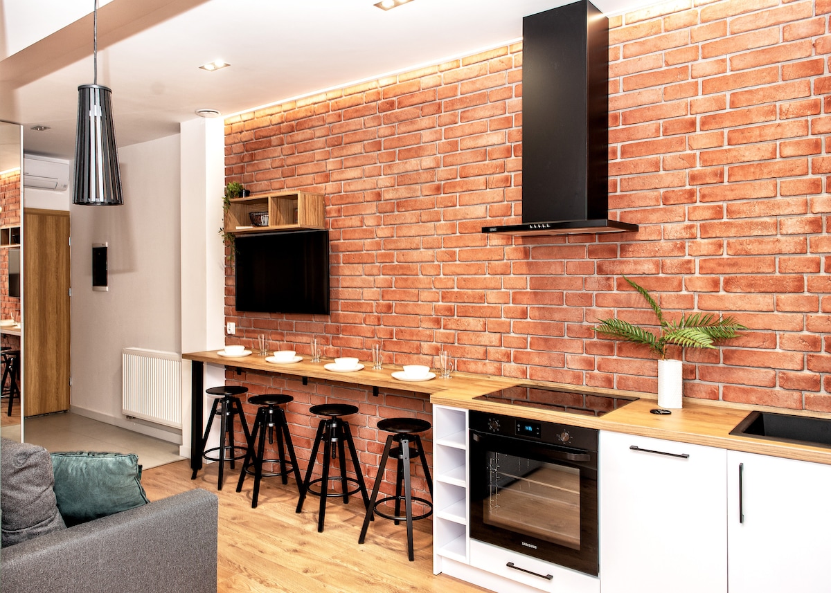 A modern kitchen features a vibrant brick wall, complemented by wooden accents and white cabinetry. Bar stools are positioned at the counter, alongside a sleek induction stove and built-in oven. A small plant adds a touch of greenery to the space.