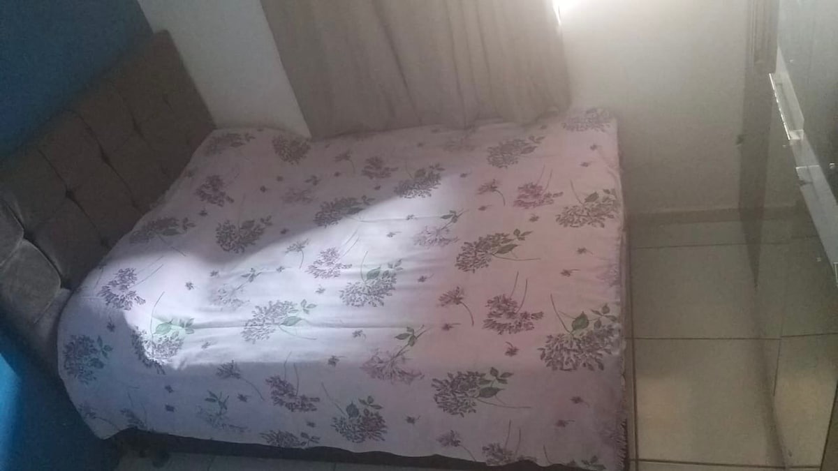 A separate sleeping area features a spacious bed covered with a floral-patterned blanket. Soft natural light filters through a window covered with light curtains, enhancing the room's calming ambiance. A simple wall and a mirror are visible, contributing to the room's functional design.