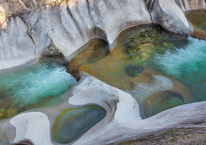 Clear turquoise waters flow over smooth, pale rocks, creating natural pools and gentle ripples. The water's vibrant hues contrast with the light-colored stone, which is shaped by years of erosion, contributing to an organic and serene landscape.