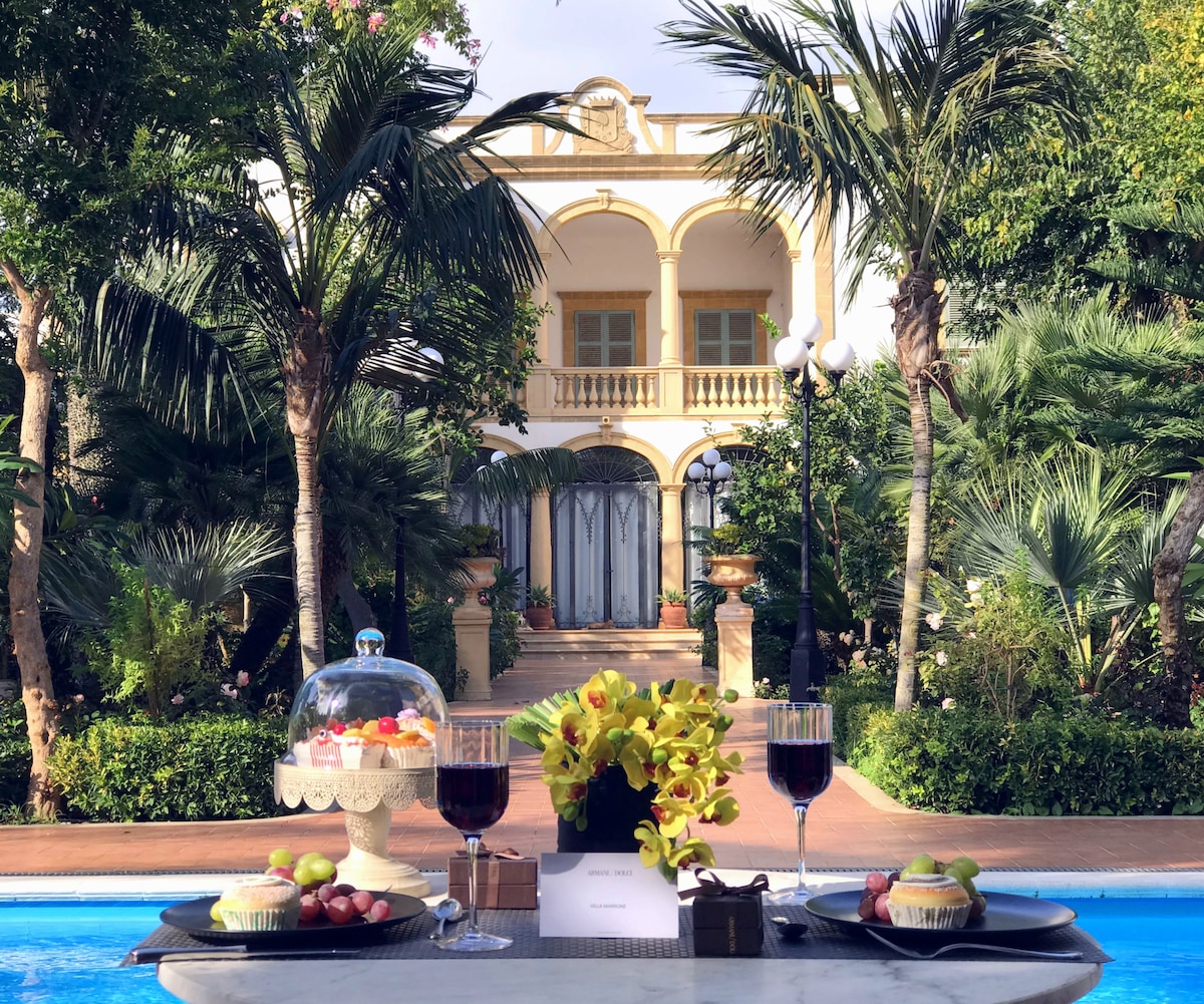 A scenic view showcases a charming villa framed by lush palm trees and vibrant greenery. A table is elegantly set with two glasses of red wine and a decorative cake stand adorned with colorful sweets, inviting a relaxing outdoor experience.