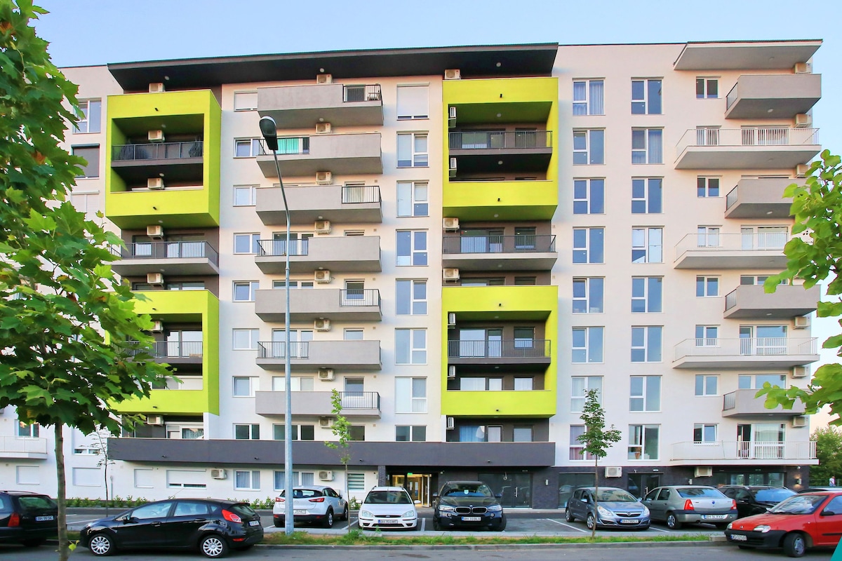A modern residential complex is displayed, featuring a combination of neutral and vibrant yellow accents. Multiple floors are visible, with balconies and air conditioning units adorning the exterior. A private parking area is accessible at the front, completed by a row of parked cars.
