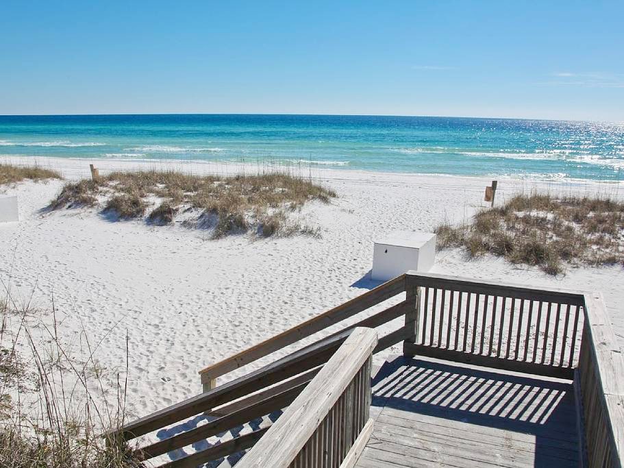 A wooden boardwalk leads to a serene beachfront, featuring soft white sand and gentle turquoise waves. Grassy patches are visible along the shore, under a clear blue sky that enhances the tranquil coastal view.
