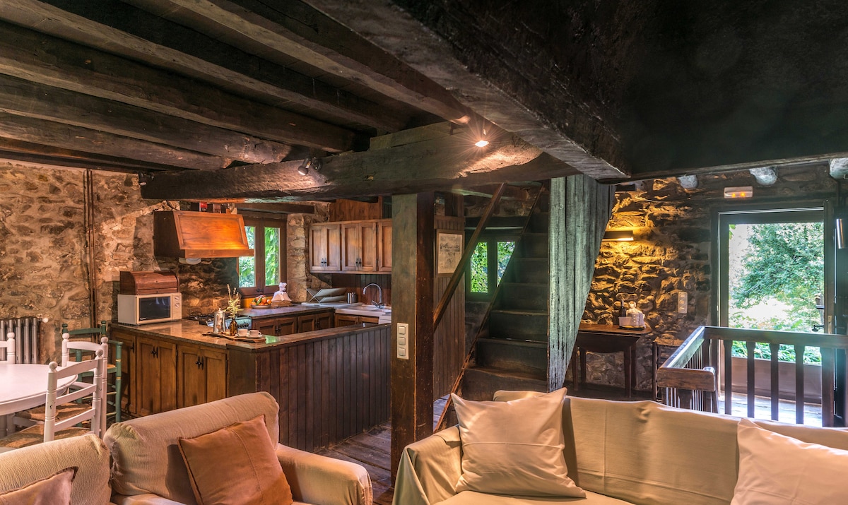 An inviting living area, featuring exposed wooden beams and stone walls. A spacious kitchen with wooden cabinetry is seen in the background. A staircase leads to the upper level, while a comfortable sofa invites relaxation. Natural light filters in through the nearby windows.