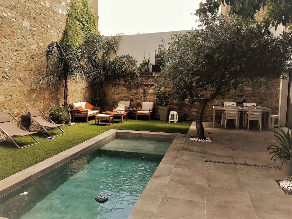 A private garden features a turquoise pool surrounded by natural stone tiles. Sun loungers and comfortable seating areas provide relaxation options, while a dining table accommodates outdoor meals. Lush greenery, including palm trees and an olive tree, enhances the serene atmosphere.