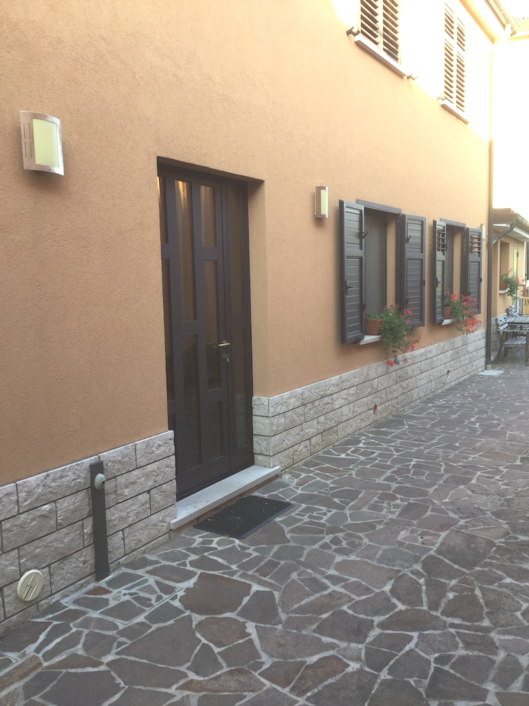An exterior view of a charming house features a prominent entrance door framed by subtle light fixtures. Flanking windows with dark shutters and adorned with potted flowers create an inviting façade. A stone pathway leads to the front door, enhancing the welcoming atmosphere.