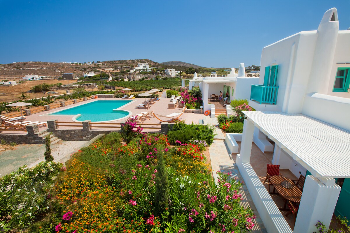 An aerial view of a charming villa complex is presented, featuring a spacious swimming pool surrounded by sun loungers and vibrant landscaping. The villa's outdoor seating area with a dining table is visible, along with colorful flowers providing a lush contrast to the white architecture. Clear sky enhances the inviting ambiance.