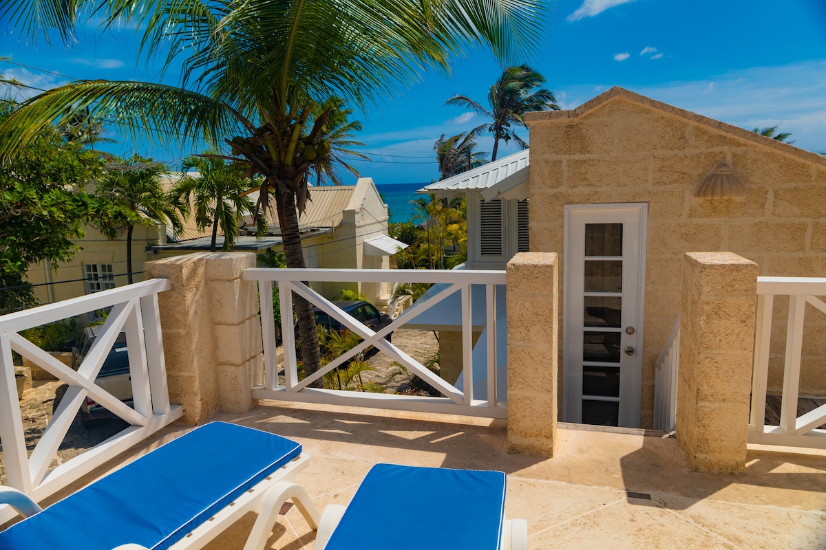 A terrace area featuring two blue lounge chairs offers an inviting space for relaxation. The stone wall surrounds the terrace, with a door leading into the interior. Palm trees and the ocean view complete the tropical setting, contributing to a serene atmosphere.