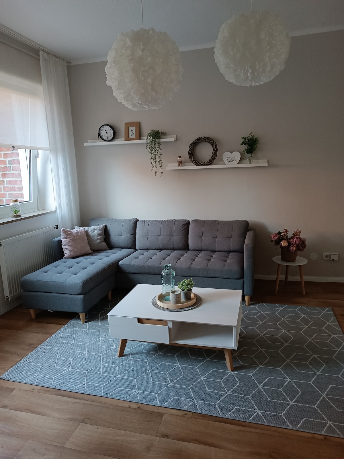 The living room features a gray sectional sofa with a chaise lounge, positioned next to a window that allows natural light. A minimalist white coffee table rests on a geometric-patterned rug, complemented by decorative plants and a small flower arrangement on the table.