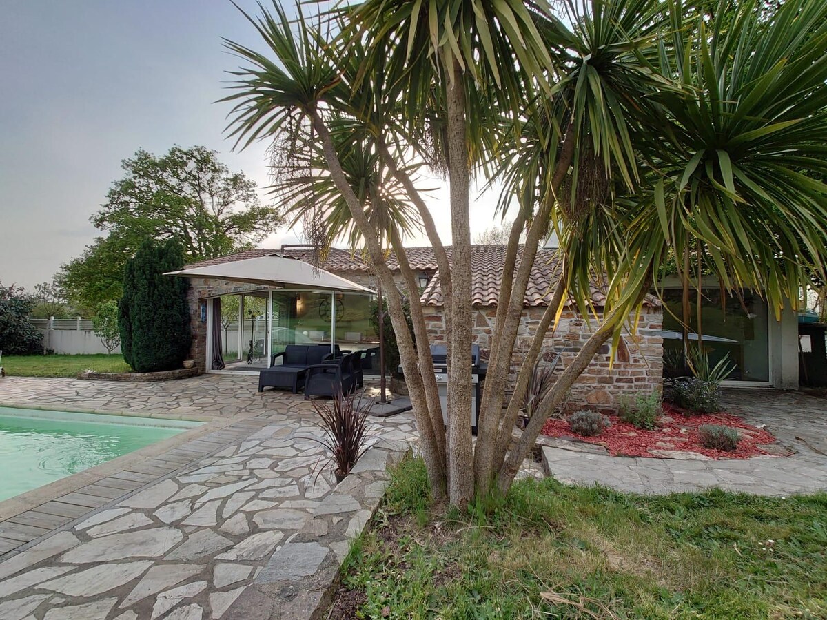 A stone cottage is surrounded by lush greenery, including palm trees, with a terrace featuring outdoor seating. A shared swimming pool is visible, complemented by a stone pathway that enhances accessibility to the outdoor space.
