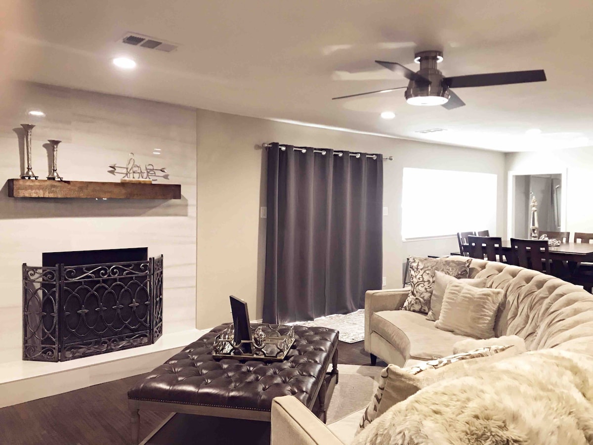 A cozy living area showcases a plush light-colored sofa and an elegant coffee table with a tufted design. A decorative fireplace with an ornate screen is flanked by minimalist decor on a wooden shelf. Privacy curtains frame a large window, allowing soft natural light to enter.