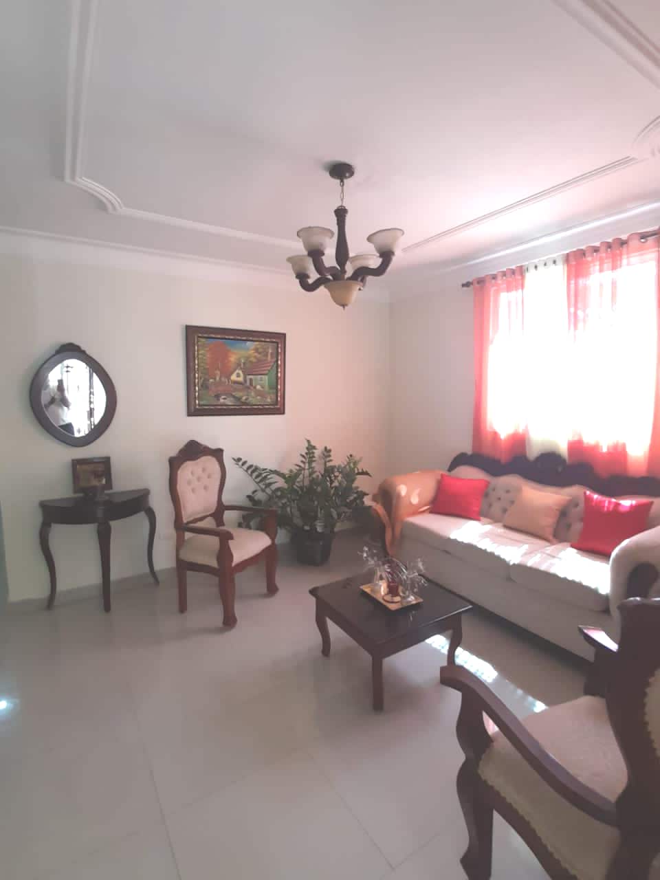 A living area features a light-colored sofa adorned with colorful cushions. Two elegant chairs are positioned nearby, alongside a small table. A decorative mirror hangs on the wall, reflecting the serene ambiance created by natural light filtering through the red-accented curtains.