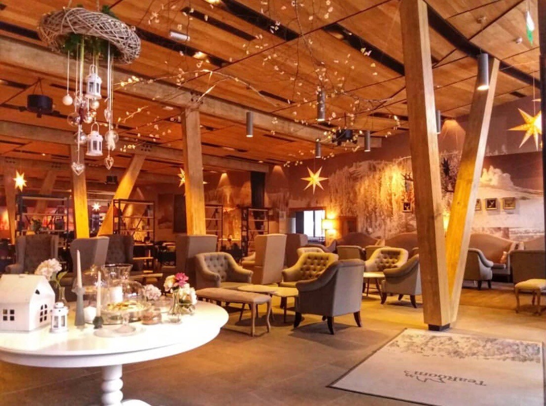 A spacious lounge area is filled with comfortable seating arrangements featuring soft armchairs and tables. Rustic wooden beams extend across the ceiling, while warm lighting creates a welcoming ambiance. Decorative elements, such as star-shaped accents and delicate floral arrangements, add charm to the relaxed setting.