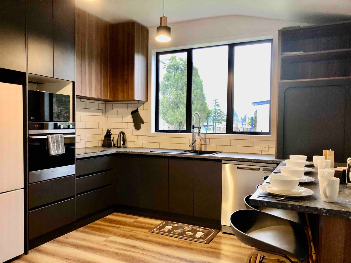 A modern kitchen is furnished with sleek black cabinetry and warm wooden accents. Natural light enters through large windows, illuminating the countertop. A dinnerware set for four is neatly arranged, and contemporary appliances, including an oven and a dishwasher, enhance the kitchen's functionality.