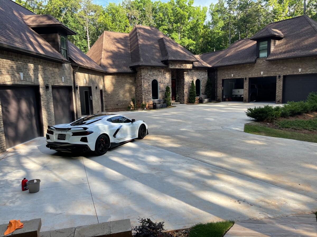 A spacious driveway is framed by a charming stone facade of the home, featuring elegant roofs and multiple garage doors. A parked vehicle is seen prominently in the foreground, with greenery surrounding the area, enhancing the serene atmosphere of the property.