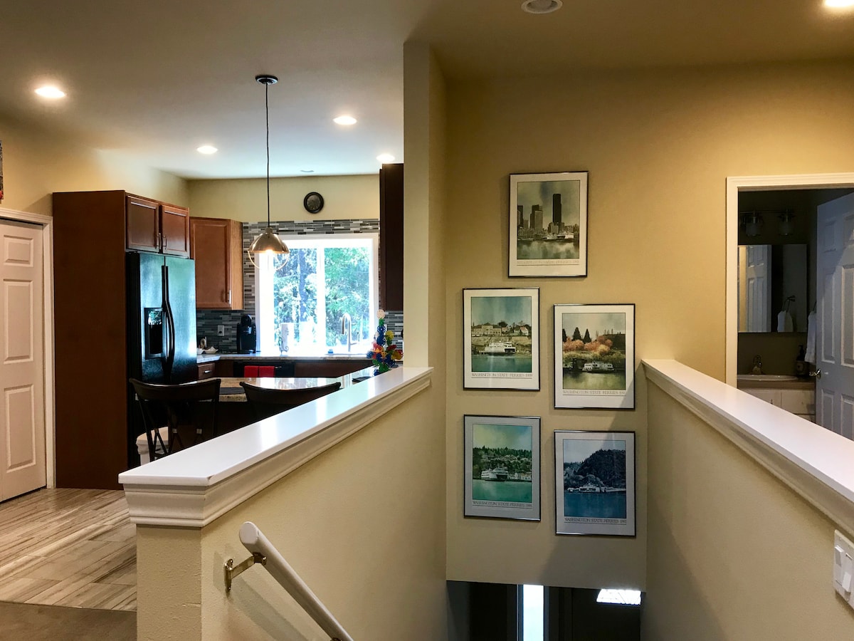 A spacious entryway is shown from an elevated angle, featuring a light-colored staircase and a landing area adorned with framed artworks. The open-concept kitchen is visible in the background, furnished with wooden cabinetry and a countertop, illuminated by natural light from nearby windows.