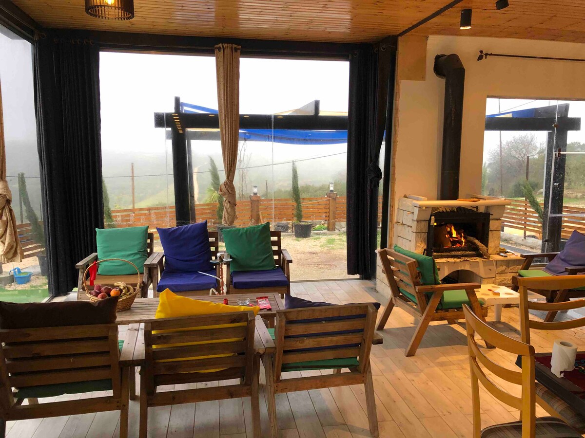 An open living area features comfortable seating, including colorful chairs and a rustic wooden table. A fireplace is positioned against one wall, providing a cozy focal point. Large windows present views of the outdoor landscape, allowing natural light to fill the space.