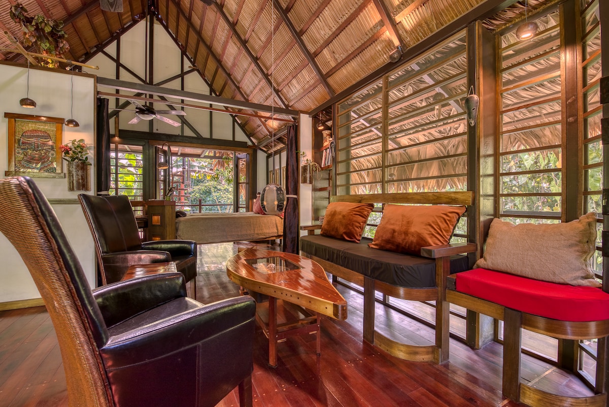 An open interior space features a combination of comfortable seating, including black leather chairs and a cozy bench with plush cushions. Large windows with screens enhance the natural light. A decorative wooden table adds a touch of elegance to the warm wooden floors.