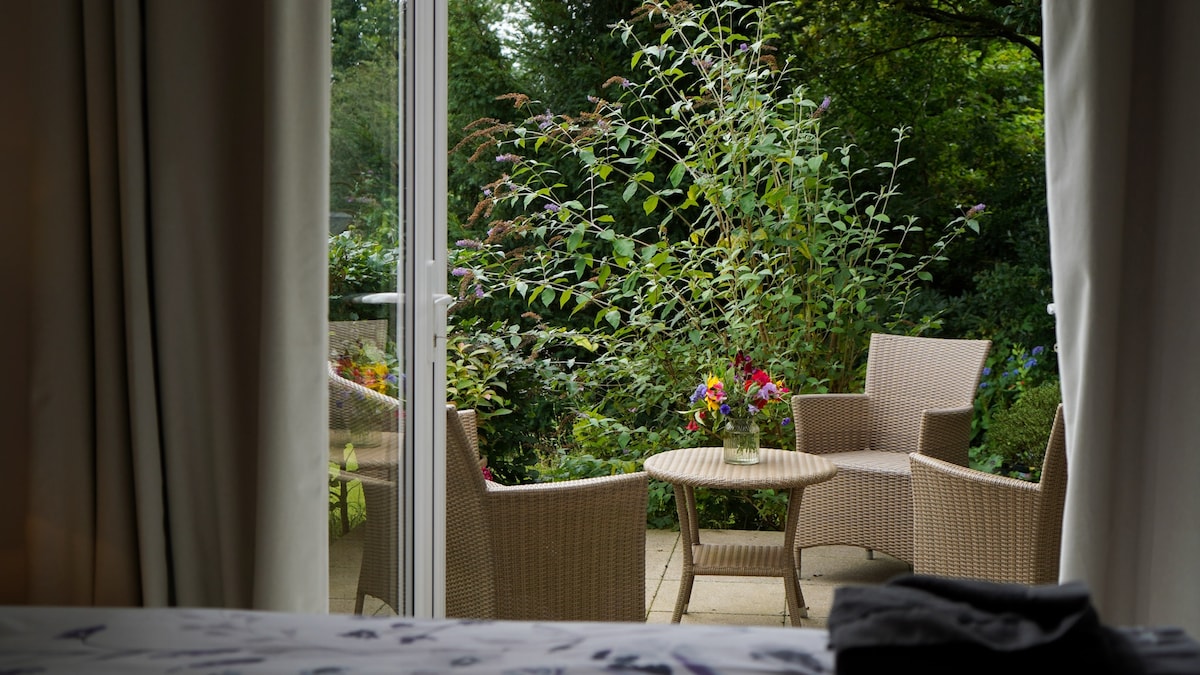A private patio is showcased through glass doors, revealing two woven chairs and a small round table surrounded by lush greenery and colorful flowers. Natural light filters in, creating a serene and inviting outdoor space.