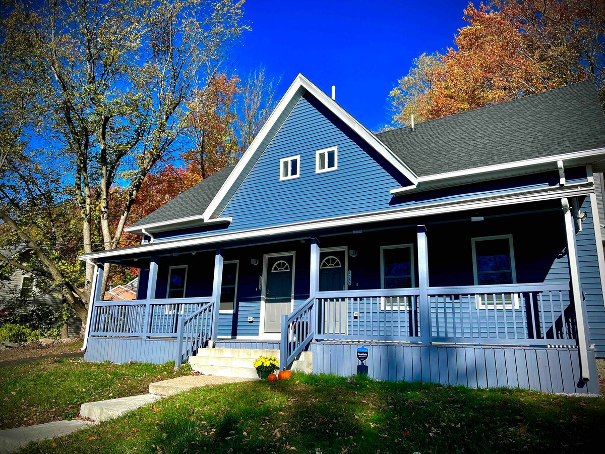 A blue-clad duplex is displayed, featuring a welcoming front porch with a rail, two entrance doors, and decorative windows. Surrounding trees show seasonal foliage in vibrant autumn colors. A concrete pathway leads to the entrance, framed by lush green grass.