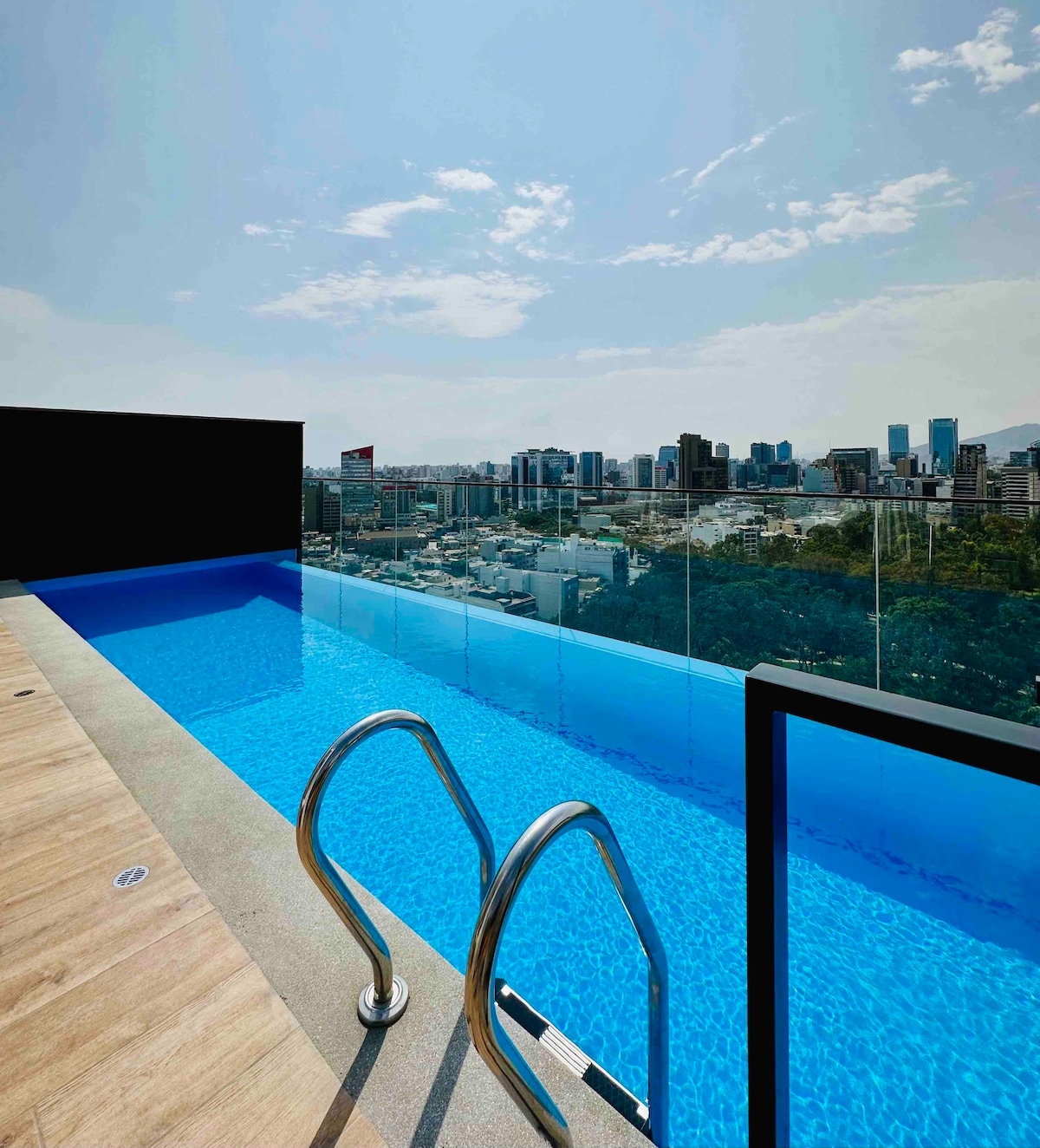 A stunning infinity pool is observed, extending towards the skyline of Lima. Clear blue water reflects the sky, with modern glass railings providing a safe, unobstructed view of the city. The pool area is spacious, featuring steps leading in for easy access.