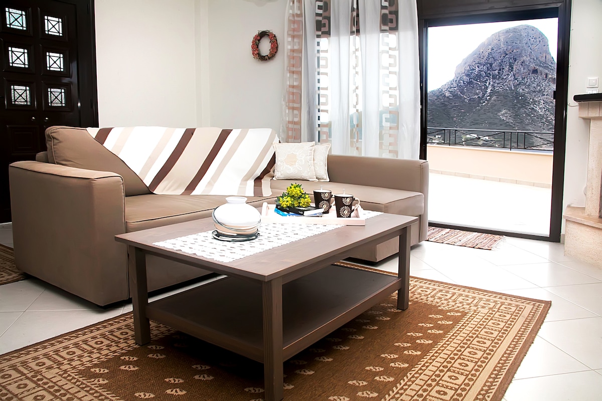 A comfortable living area is shown, featuring a beige sofa and a light brown armchair. A simple coffee table sits centrally, adorned with decorative items. Large glass doors open to a balcony, providing a clear view of the mountain landscape outside. Natural light fills the space.