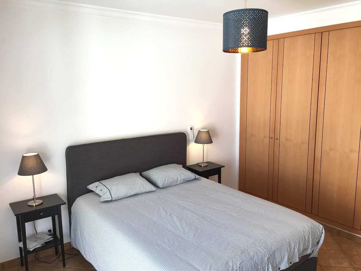 A spacious bedroom features a comfortable bed with patterned bedding, flanked by two bedside tables with lamps. A warm light fixture hangs above, illuminating the room. A large wooden wardrobe occupies one wall, providing ample storage space.