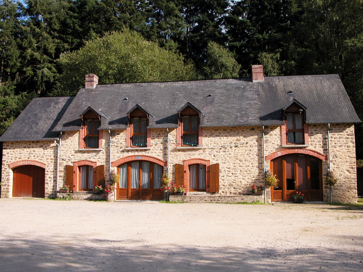 A stone cottage is presented with a symmetrical facade, featuring multiple arched windows and doors. Red brick accents complement the natural stone exterior. Flower pots are positioned at the base of the windows, bringing a touch of greenery to the entrance.