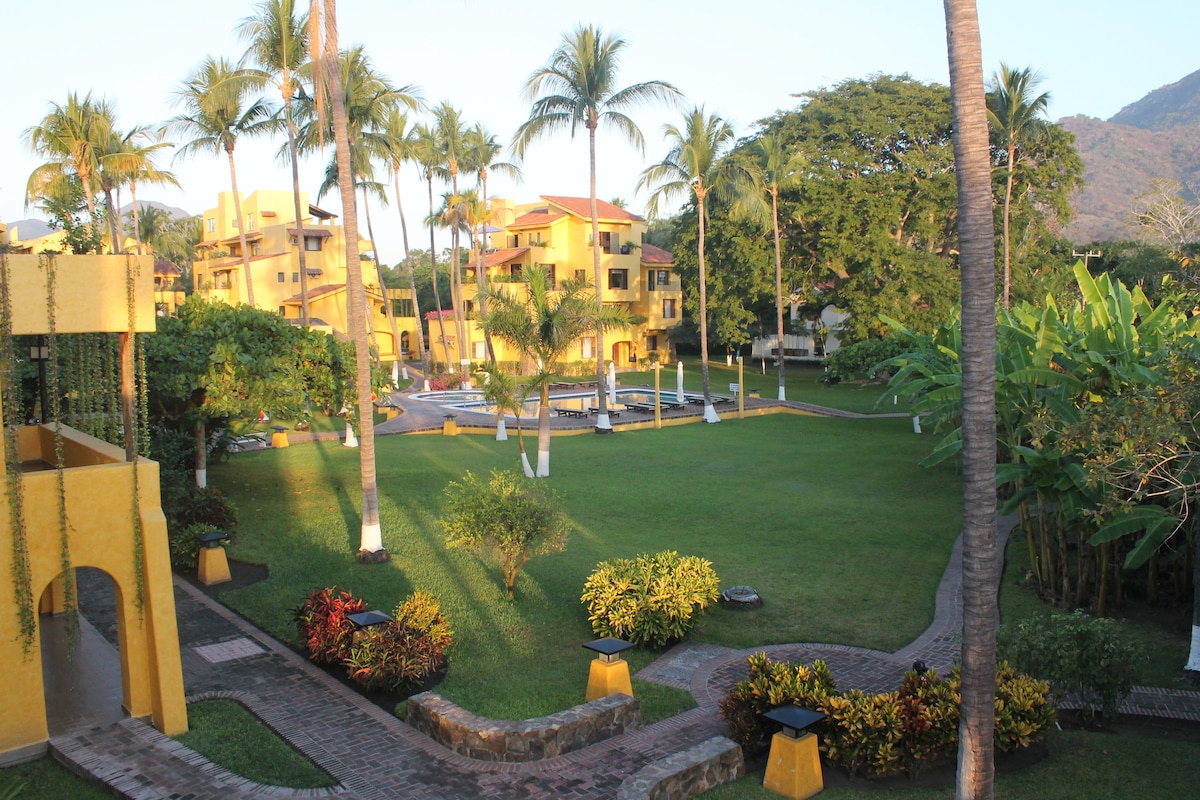 A well-maintained green area is surrounded by palm trees and colorful landscaping. Warm yellow buildings form a picturesque backdrop, while a pathway meanders through the space. The serene environment includes a swimming pool visible in the distance, evoking a tranquil atmosphere for relaxation.