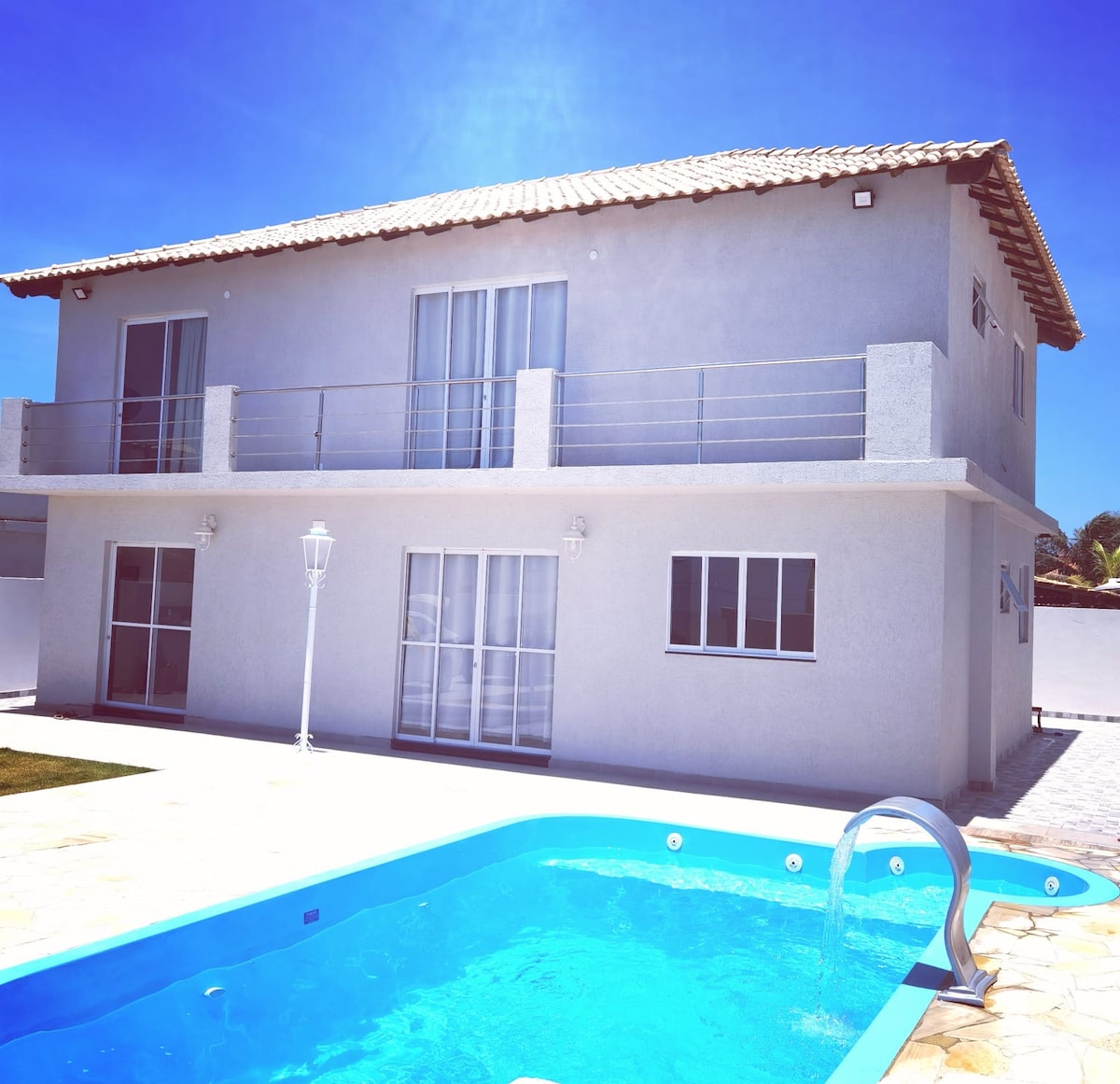 A modern two-story house is depicted, featuring large glass doors and balconies on the upper floor. A swimming pool is positioned in the foreground, surrounded by natural stone decking. The exterior exhibits a clean, neutral color palette under a clear blue sky.