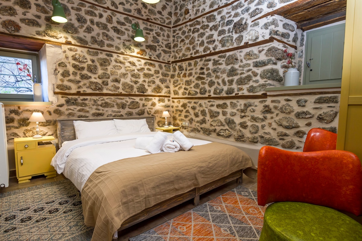 A cozy sleeping area features a spacious bed with neutral bedding and white towels arranged neatly. The natural stone walls are complemented by vibrant, uniquely shaped chairs and warm lighting from bedside lamps, creating a welcoming ambiance.