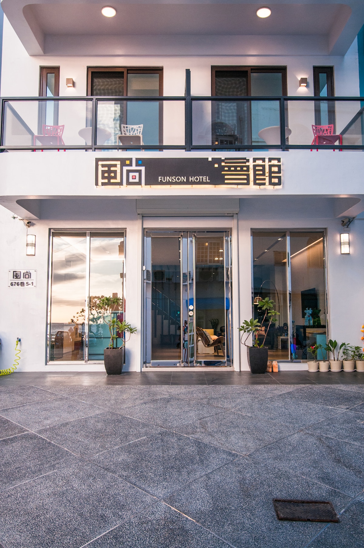 The exterior of Funson Hotel features modern architecture with large glass doors and a welcoming entrance. Potted plants flank the entrance, and there are colorful chairs on the balcony above. The surface is tiled, providing a sleek pathway leading into the hotel.