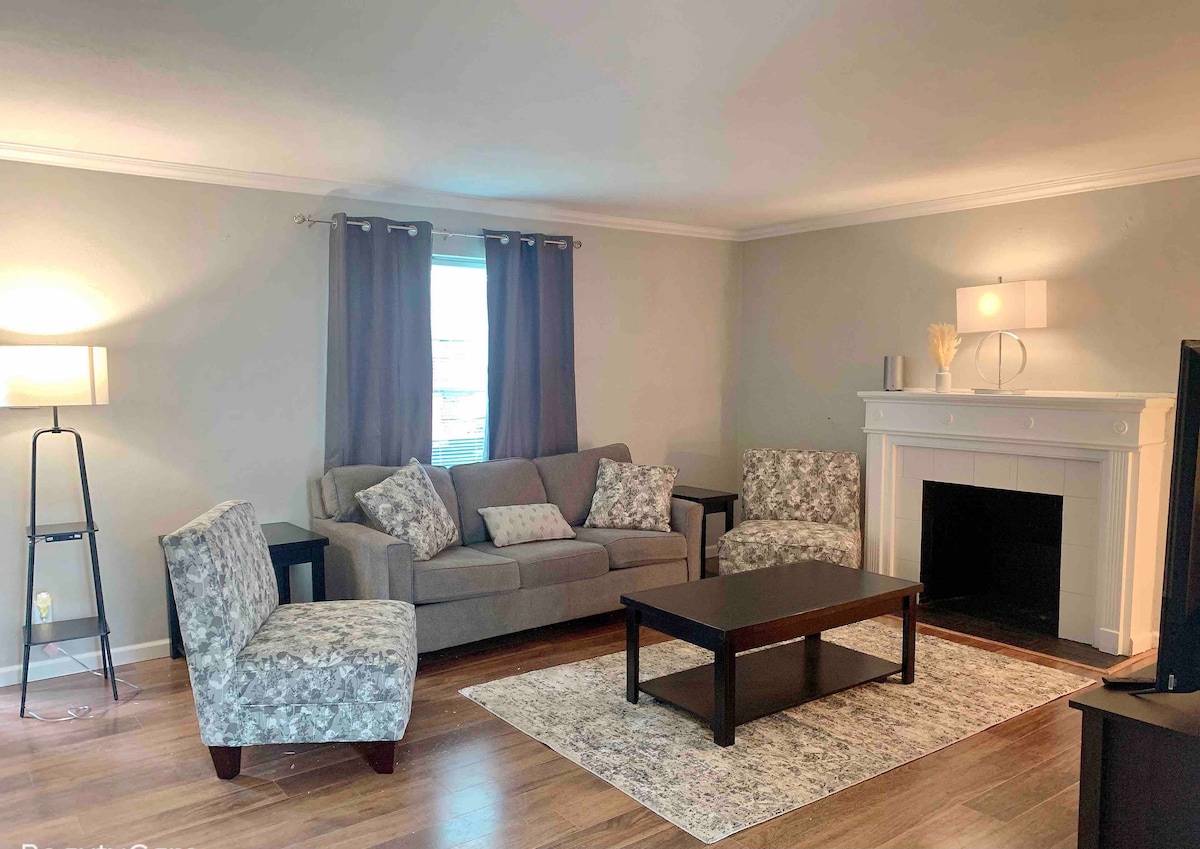 A cozy living room features a gray sofa with patterned accent chairs. A dark wooden coffee table is centered on a light area rug, while a fireplace with a simple mantle adds warmth. Natural light enters through a window, complemented by dark curtains.