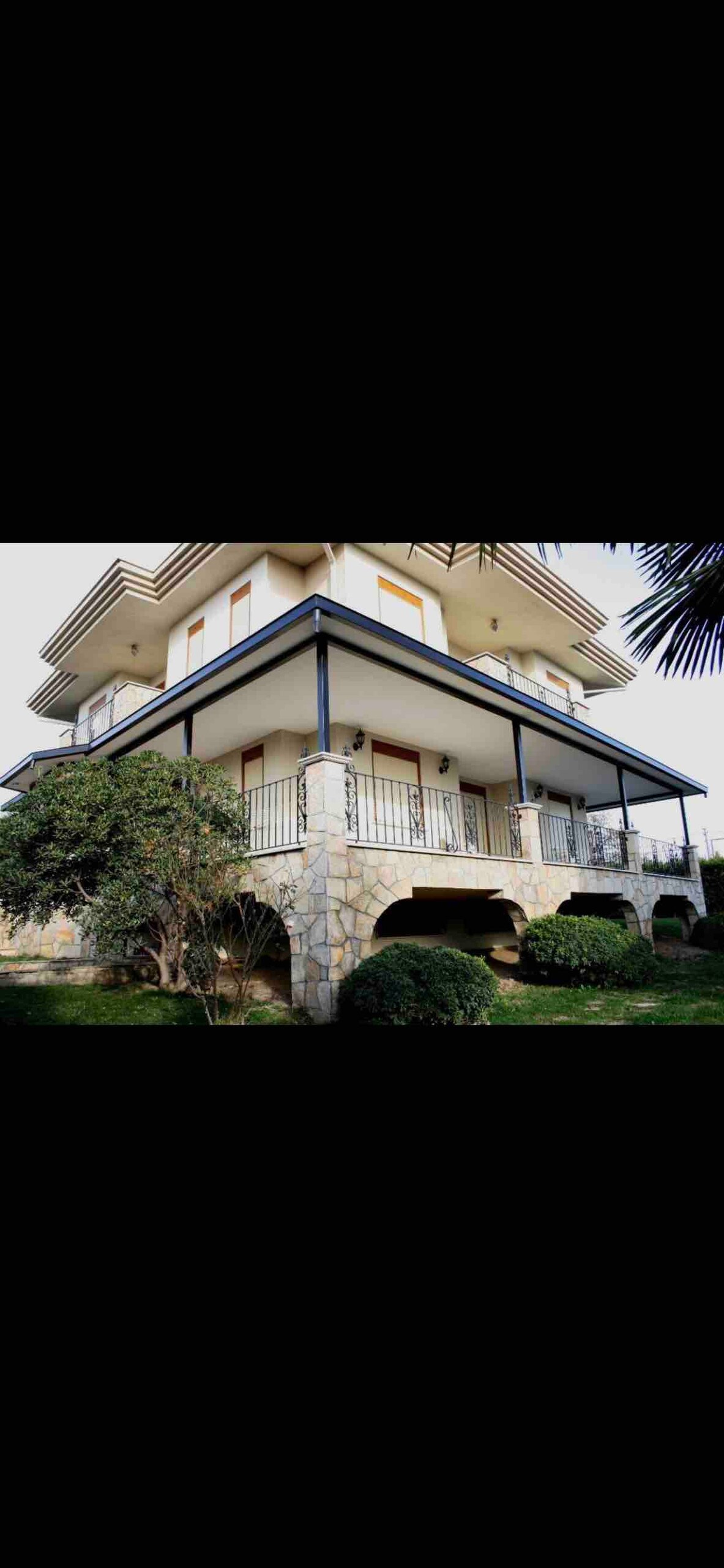 A three-story house is set within a green landscape, featuring a stone base and white exterior. The balconies are framed with black railings, while large windows allow natural light to fill the interior. Lush shrubs and trees surround the property.