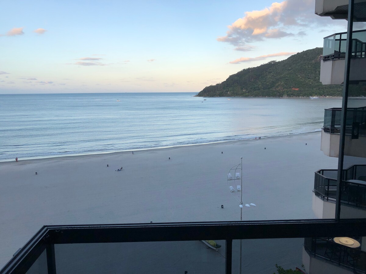A wide view of the ocean is visible from the balcony, showcasing calm waters reflecting the sky. A sandy beach stretches out, with distant figures enjoying the shoreline. Lush green hills rise in the background under a partly cloudy sky.
