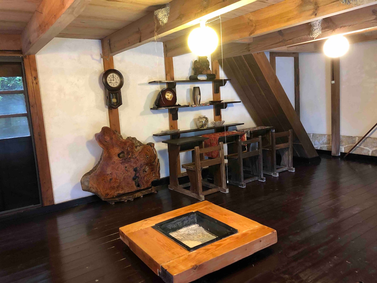 An interior space showcases traditional wooden beams and dark wooden flooring. A low wooden table serves as a centerpiece, accompanied by a small dining set against a wall adorned with decorative items. A wall clock adds a touch of nostalgia to the rustic atmosphere.