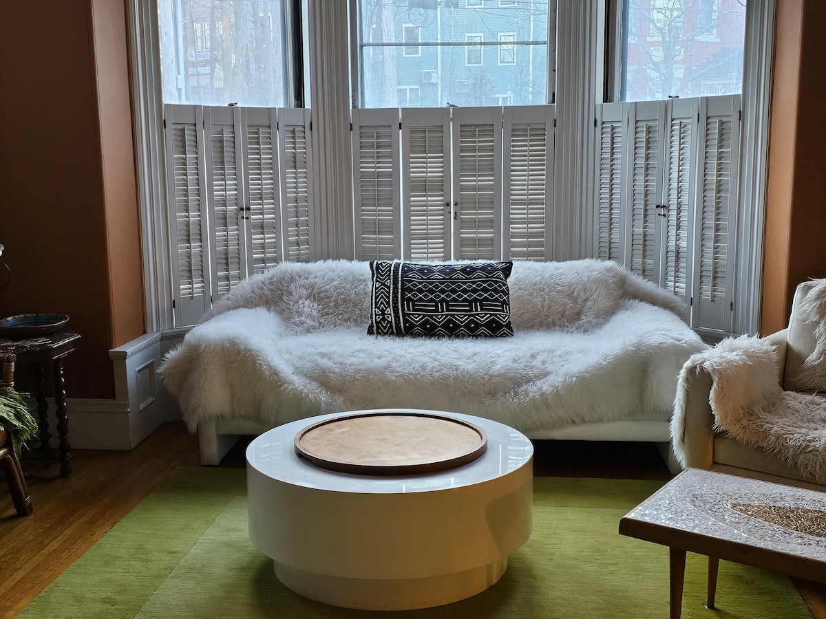 A bright seating area features a soft, white couch adorned with a patterned cushion. A circular coffee table sits at the center, complemented by a plush green area rug. Large bay windows with wooden shutters allow natural light to fill the space.