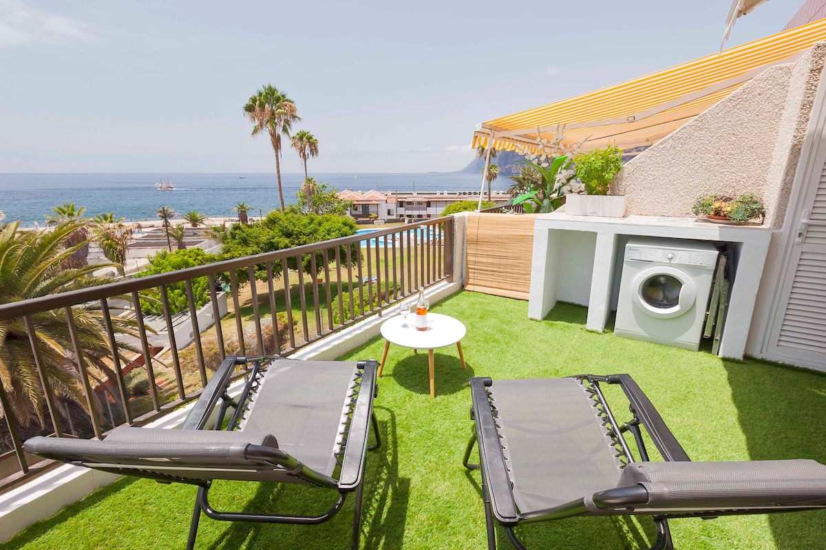 A private balcony features two lounge chairs positioned on green turf. A small round table rests between them, while a washing machine is integrated into the built-in cabinetry. The sea and palm trees are visible in the background, under a clear blue sky.