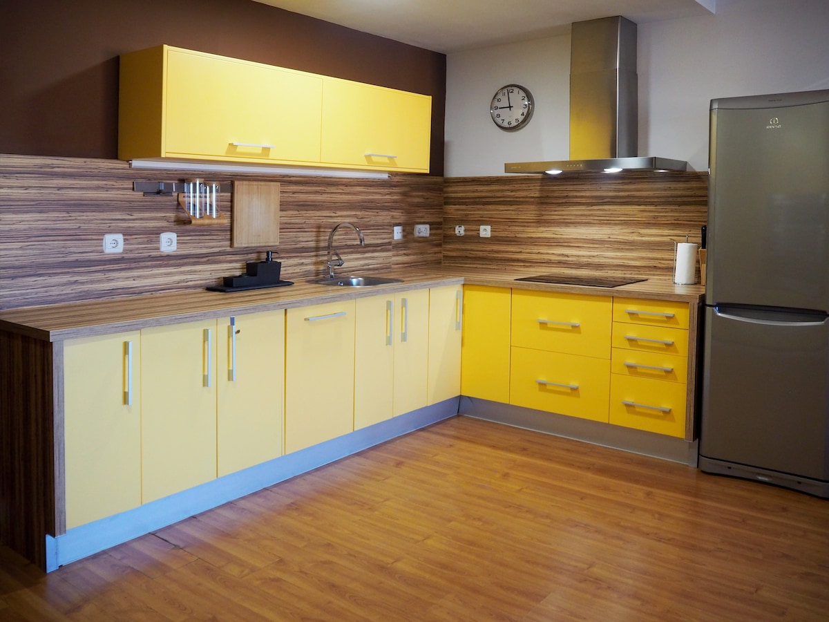 A modern kitchen is equipped with vibrant yellow cabinetry and a sleek stainless steel refrigerator. The wooden wall paneling adds warmth, while the stainless steel sink and range hood contribute to a functional layout. A clock is mounted on the wall for convenient timekeeping.