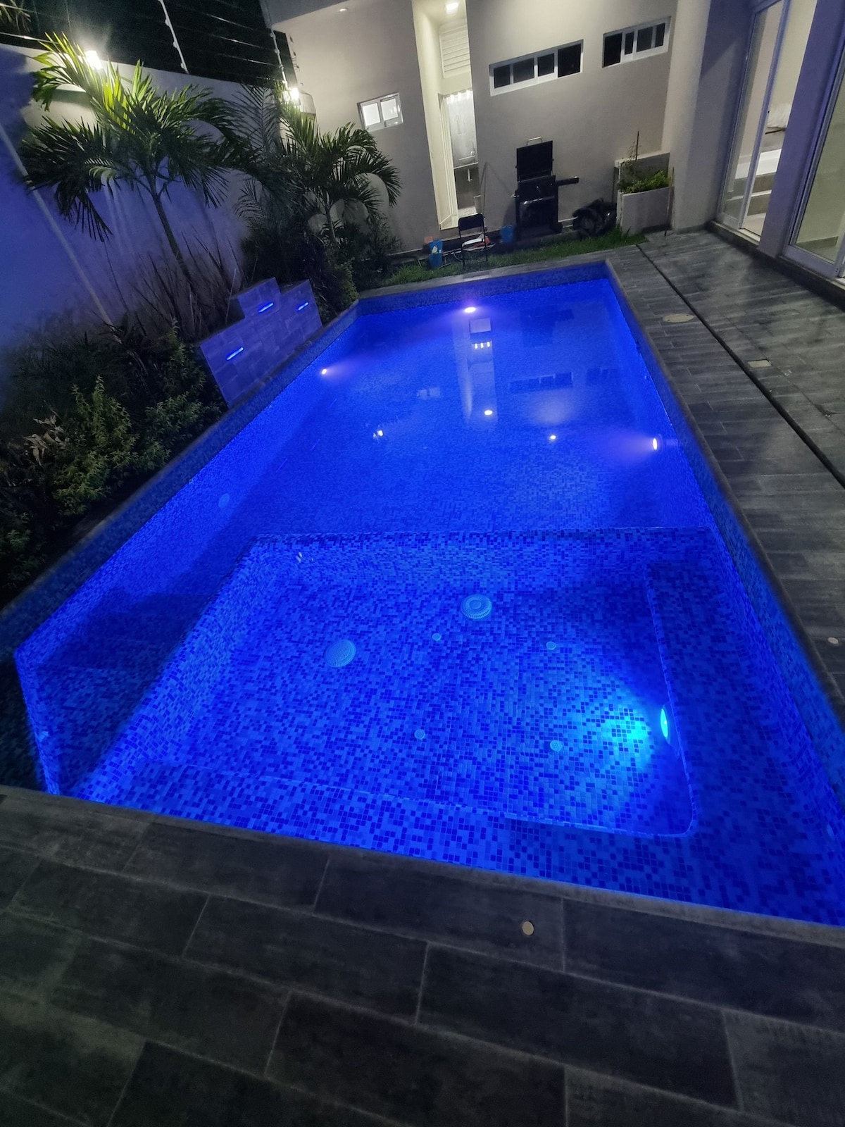 A clear swimming pool glows in vibrant blue, surrounded by textured stone tiles. The area features tropical greenery and is illuminated by subtle lighting. The jacuzzi is integrated within the pool, enhancing the relaxing ambiance of the outdoor space.