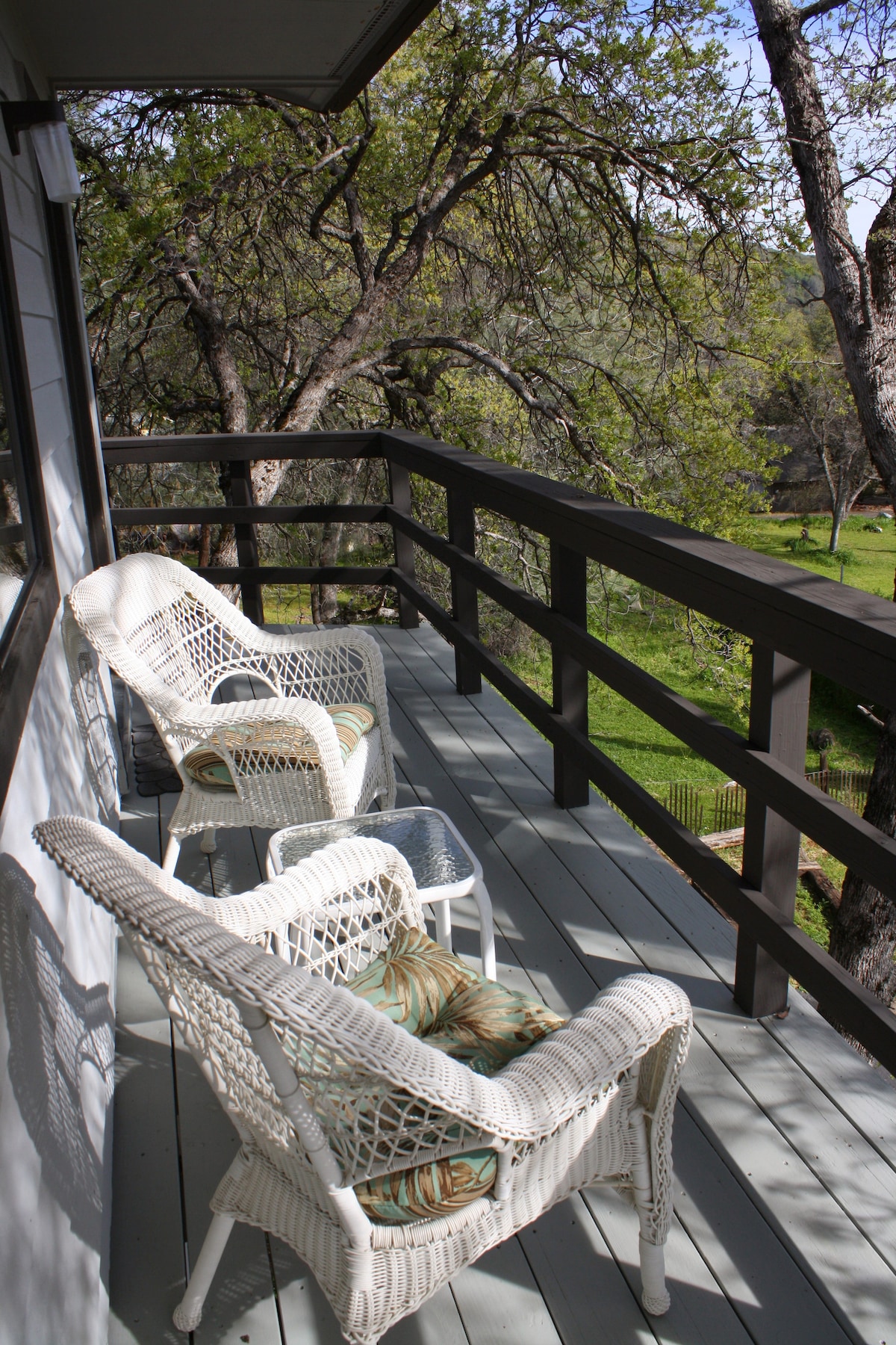 Two wicker chairs with soft cushions are positioned on a private balcony, surrounded by lush trees and a view of the green landscape below. The railing offers a sense of separation from the outdoors while enhancing the serene atmosphere.