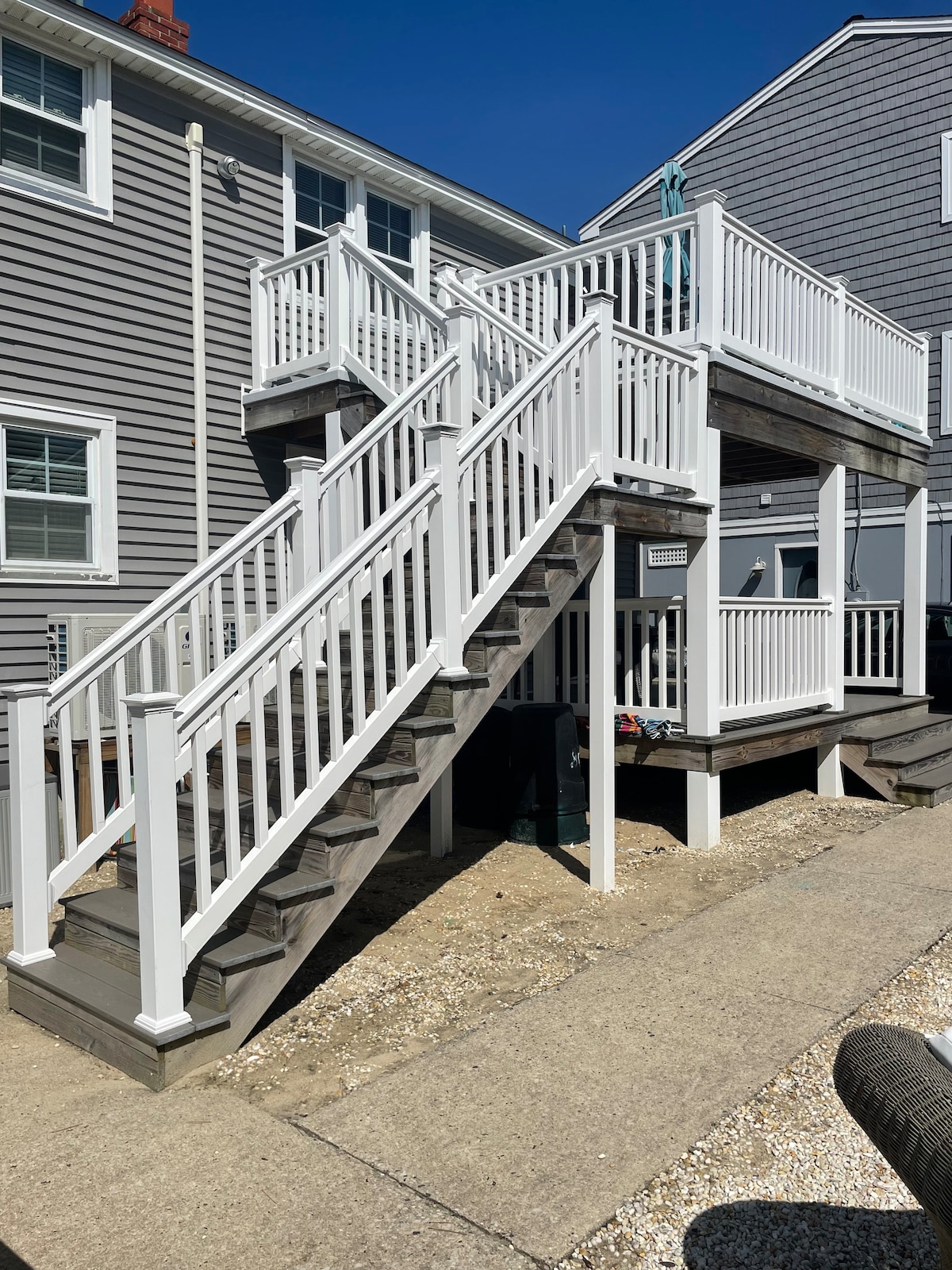 A set of white wooden stairs leads to the entrance of the second floor, flanked by railings on both sides. The surrounding area features a concrete pathway and landscaping with pebbles, creating an inviting entry to the beach house.